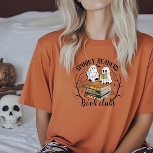 May include: An orange t-shirt with a Halloween-themed graphic. The design features two ghost characters reading books, with the text "Spooky Readers Book Clubs" in a circular arrangement. The shirt is paired with black and white patterned pants.