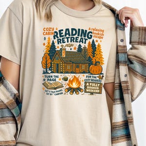 May include: Beige t-shirt with a graphic design featuring a cabin, trees, and a pumpkin, with the words "Cozy Cabin Reading Retreat" and other text related to reading and a campfire. The shirt has a crew neck and short sleeves.