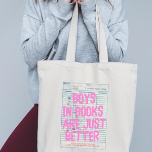 May include: Cream-colored tote bag with the text "BOYS IN BOOKS ARE JUST BETTER" in pink. The text is superimposed over a library card design. The bag has two cream-colored handles.