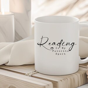 May include: White ceramic mug with the text "Reading Is My Favorite Sport" in black script. The mug has a handle and is sitting on a wooden surface. Candles and fabric are in the background.