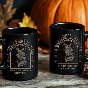 May include: Two black ceramic mugs with the text "A WELL-READ WOMAN IS A DANGEROUS CREATURE" in gold lettering. The design includes an open book, flowers, and celestial elements. The mugs are on a wooden surface with a pumpkin in the background.