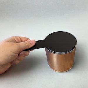 May include: A copper-colored cylindrical container with a black lid and handle. The container is held by a person's hand. The lid is round and covers the top of the container.