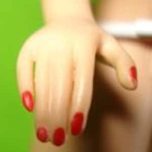 May include: Close-up of a doll's hand with red nail polish. The hand is slightly curved, with fingers extended. The skin tone is light, and the background is a solid green.