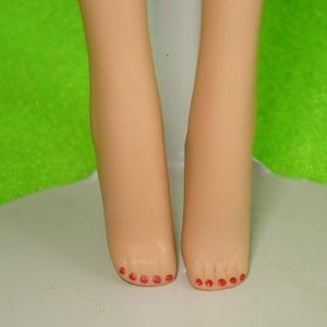May include: A close-up of two doll legs with painted red toenails. The legs are a light tan color and are positioned close together. The background is a mix of white and green.