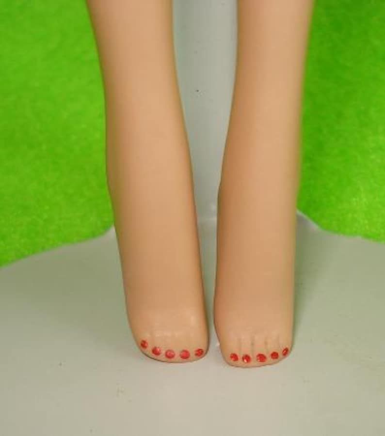 May include: A close-up of two doll legs with painted red toenails. The legs are a light tan color and are positioned close together. The background is a mix of white and green.