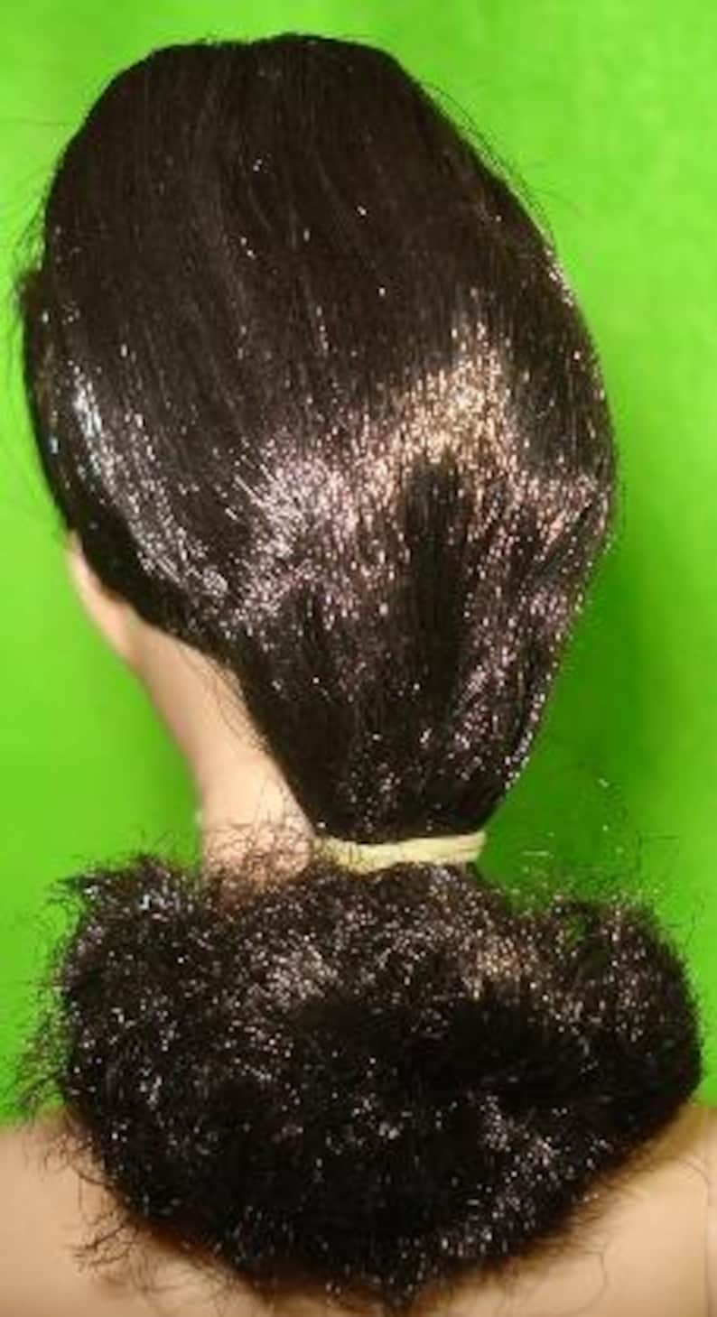 May include: A close-up of a doll's head with dark brown hair styled in a bun. The hair is pulled back and secured with a light-colored elastic band. The bun is positioned at the nape of the neck.