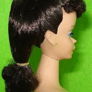 May include: A side profile of a vintage doll's head. The doll has black hair styled in a ponytail and a small bun. The doll's face is pale with blue eyes and a light pink lip color. The background is a solid green.