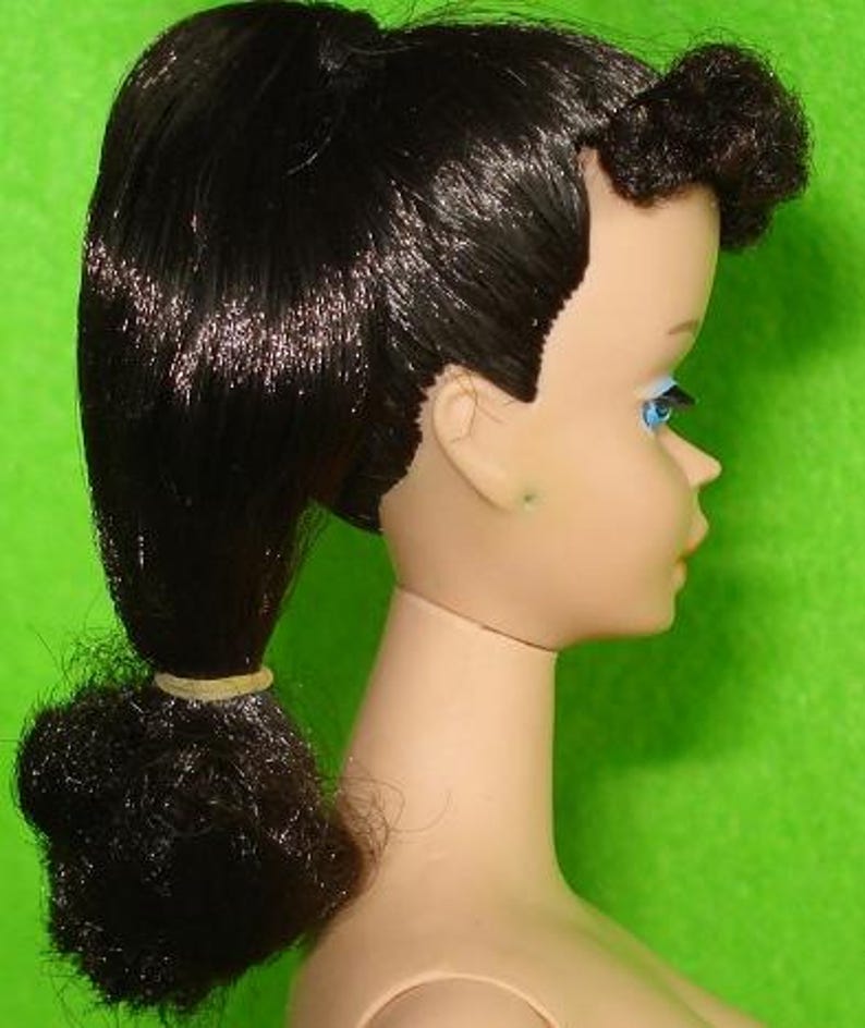 May include: A side profile of a vintage doll's head. The doll has black hair styled in a ponytail and a small bun. The doll's face is pale with blue eyes and a light pink lip color. The background is a solid green.