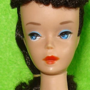 May include: Close-up of a vintage doll's face. The doll has blue eyes with black eyeliner, red lipstick, and dark hair styled in a bouffant. The doll's skin tone is fair, and the background is a solid green.