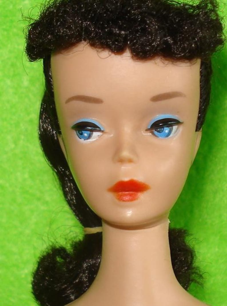 May include: Close-up of a vintage doll's face. The doll has blue eyes with black eyeliner, red lipstick, and dark hair styled in a bouffant. The doll's skin tone is fair, and the background is a solid green.