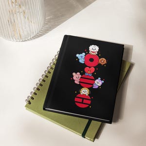 Puede incluir: Cuaderno negro con un cuaderno verde encuadernado en espiral debajo. El cuaderno negro presenta un gráfico colorido de personajes de dibujos animados dispuestos alrededor de un círculo y un corazón rojos. Los cuadernos están sobre una superficie blanca.