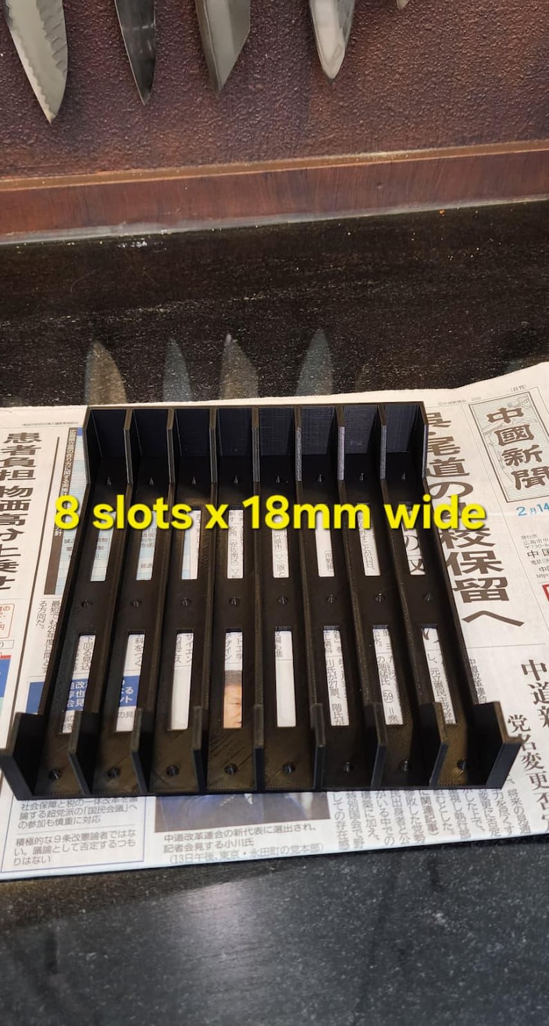 Może przedstawiać: Czarny uchwyt na noże z 8 szczelinami, każda o szerokości 18 mm, na noże kuchenne. Uchwyt jest wykonany z ciemnego materiału i został zaprojektowany tak, aby bezpiecznie trzymać noże. Tekst na uchwycie brzmi "8 slots x 18mm wide".