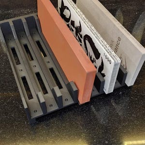 May include: A black plastic knife sharpening stone holder with three sharpening stones. The stones are a reddish-brown, white with black text, and light gray. Knives are visible in the background.
