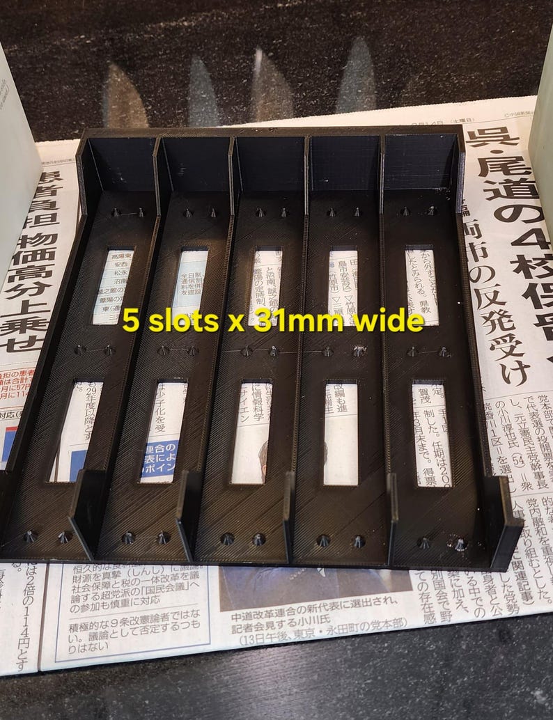 Może przedstawiać: Prezentowany jest czarny plastikowy organizer z pięcioma szczelinami, każda o szerokości 3,1 cm. Organizer ma prostokątny kształt z pionowymi przegrodami. Widoczny jest tekst "5 slots x 31mm wide".