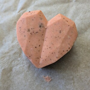 Puede incluir: Una bomba de baño en forma de corazón de color melocotón con un diseño geométrico. La bomba de baño tiene pequeñas motas oscuras y se encuentra sobre una superficie clara y texturizada. Un pequeño trozo de la bomba de baño está suelto.