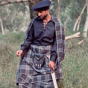 Peut inclure: Une personne portant une tenue &eacute;cossaise traditionnelle, comprenant un b&eacute;ret bleu fonc&eacute;, une chemise noire &agrave; lacets et un kilt &agrave; carreaux gris et noir. Un ch&acirc;le &agrave; carreaux assorti est drap&eacute; sur les &eacute;paules. Une &eacute;p&eacute;e et un sporran en cuir avec un embl&egrave;me de cerf sont &eacute;galement visibles.