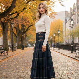 May include: A woman wearing a white turtleneck sweater and a long, pleated, navy blue plaid skirt with brown leather buckle details. She is wearing brown boots. The background shows a tree-lined street with autumn foliage.