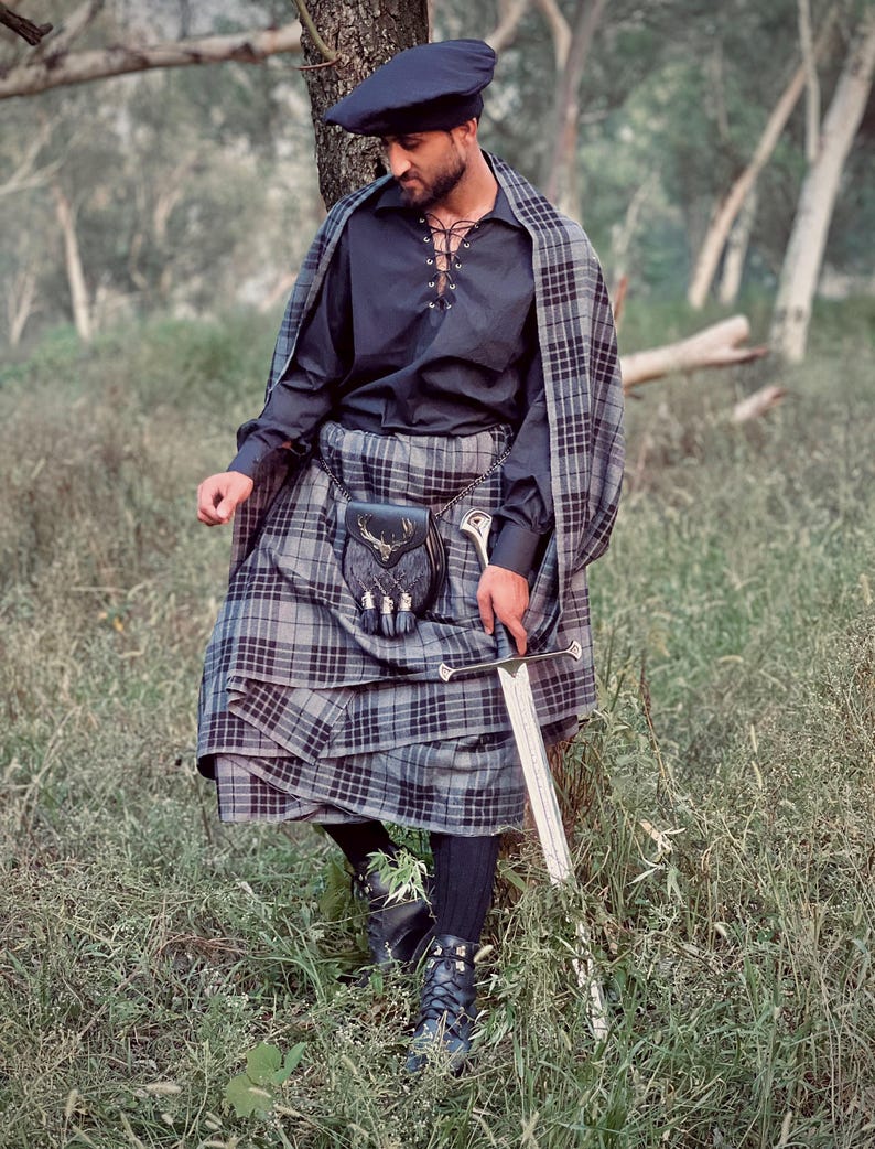 Peut inclure: Une personne portant une tenue &eacute;cossaise traditionnelle, comprenant un b&eacute;ret bleu fonc&eacute;, une chemise noire &agrave; lacets et un kilt &agrave; carreaux gris et noir. Un ch&acirc;le &agrave; carreaux assorti est drap&eacute; sur les &eacute;paules. Une &eacute;p&eacute;e et un sporran en cuir avec un embl&egrave;me de cerf sont &eacute;galement visibles.