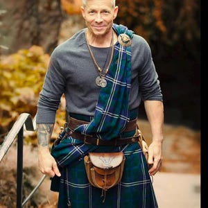 Può includere: Un uomo con un kilt scozzese tradizionale con un motivo a quadri blu e verdi. Indossa anche una maglietta grigia a maniche lunghe, una cintura di cuoio marrone e uno sporran di cuoio. Una sciarpa a quadri è drappeggiata sulla spalla.