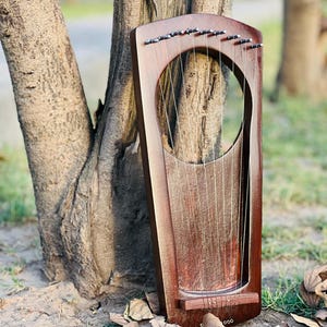 May include: A wooden lyre with a dark brown finish, featuring a circular sound hole and multiple strings. The instrument is leaning against a tree trunk, with dried leaves scattered on the ground. The lyre's design is simple and elegant.