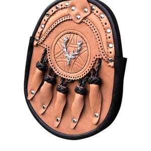 May include: A tan leather sporran with a black border and decorative silver rivets. The sporran features a central emblem with a silver stag design and dangling leather tassels with silver studs. A traditional Scottish accessory.