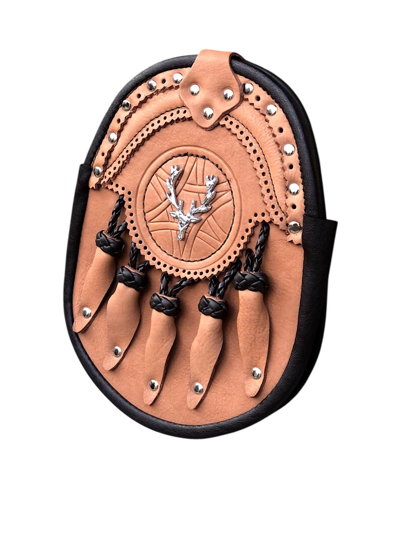 May include: A tan leather sporran with a black border and decorative silver rivets. The sporran features a central emblem with a silver stag design and dangling leather tassels with silver studs. A traditional Scottish accessory.