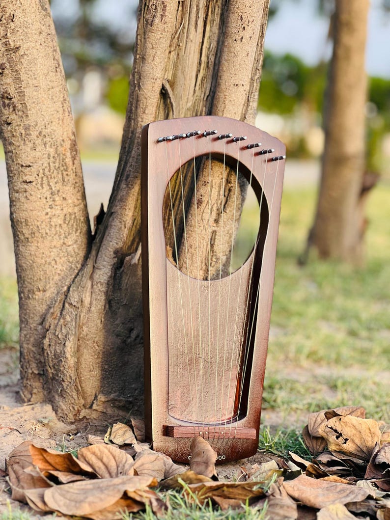 May include: A wooden lyre with strings, featuring a large, open sound hole. The instrument has a dark brown finish and is positioned outdoors near a tree trunk, with fallen leaves in the foreground. The strings are visible and taut.