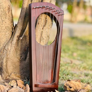 May include: A dark brown wooden lyre with strings, featuring a large circular opening. The instrument is leaning against a tree trunk, with fallen leaves scattered on the ground. The lyre's strings are visible, and the wood has a polished finish.