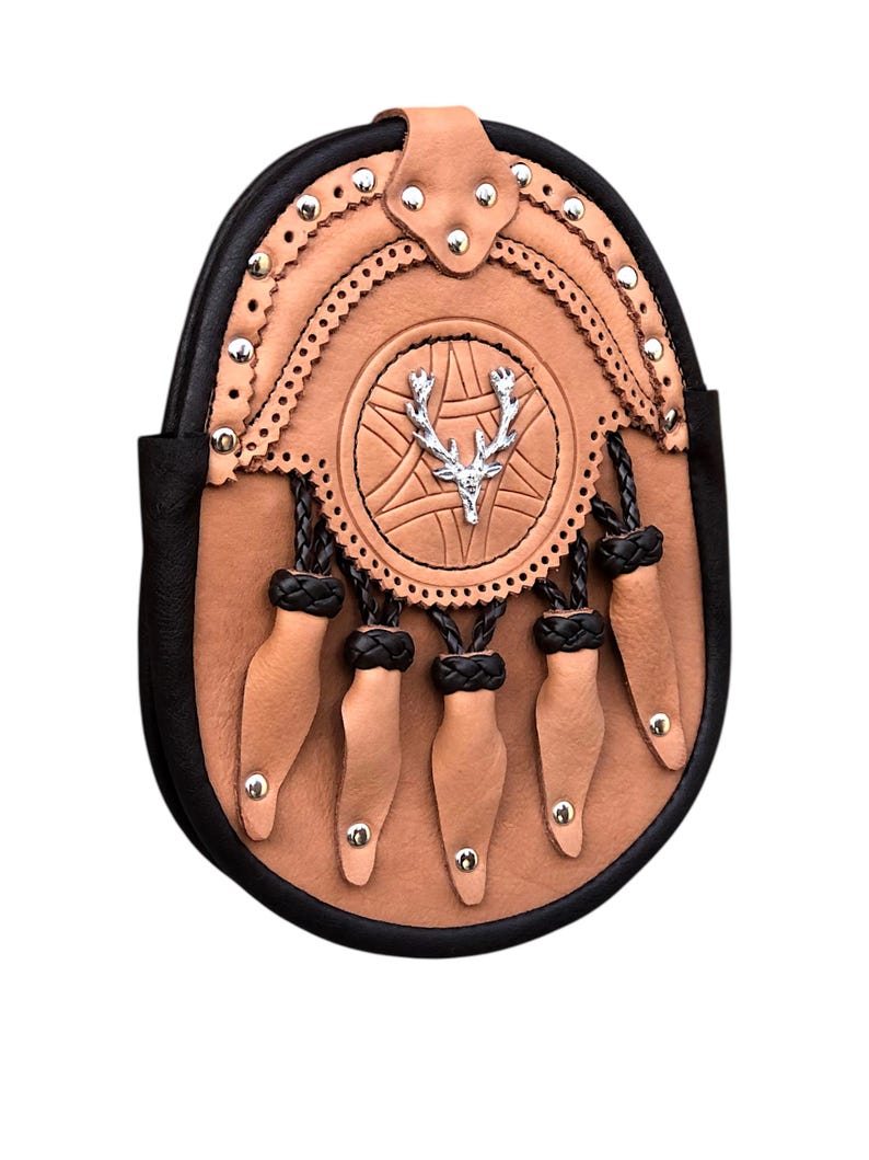 May include: A tan leather sporran with a black border and decorative elements. The sporran features a silver stag emblem in the center and five tan leather tassels hanging from the bottom. Silver studs and decorative stitching add detail.