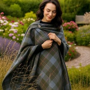 Può includere: Una donna indossa uno scialle a quadri verdi, blu e rosa con una spilla decorativa. Lo scialle è drappeggiato sulle spalle. Lo sfondo presenta un giardino con fiori e vegetazione.