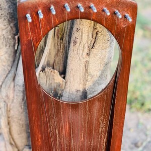 May include: A handcrafted, wooden lyre with a rich, reddish-brown finish. The instrument features a circular sound hole and multiple strings stretched across its body. The lyre is positioned against a natural, textured background, highlighting its traditional design.