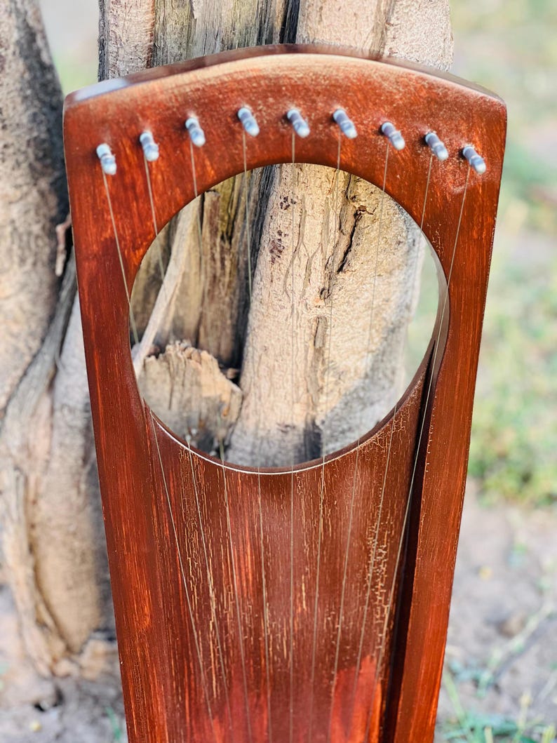 May include: A handcrafted, wooden lyre with a rich, reddish-brown finish. The instrument features a circular sound hole and multiple strings stretched across its body. The lyre is positioned against a natural, textured background, highlighting its traditional design.