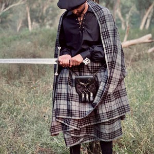 Peut inclure: Une personne portant un b&eacute;ret noir, une chemise noire avec des lacets et un kilt et une cape &agrave; carreaux gris. Un sporran noir avec des glands est suspendu &agrave; la taille. La personne tient une longue &eacute;p&eacute;e argent&eacute;e.