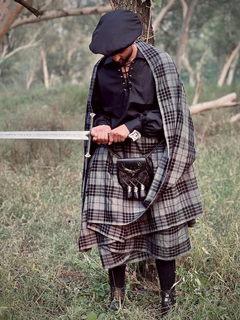 Peut inclure: Une personne portant un b&eacute;ret noir, une chemise noire avec des lacets et un kilt et une cape &agrave; carreaux gris. Un sporran noir avec des glands est suspendu &agrave; la taille. La personne tient une longue &eacute;p&eacute;e argent&eacute;e.