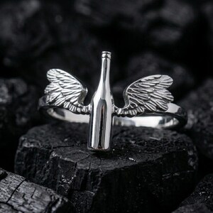 May include: A silver ring featuring a wine bottle design with detailed wings on either side. The ring is set against a dark, textured background, highlighting the metallic shine of the jewelry.