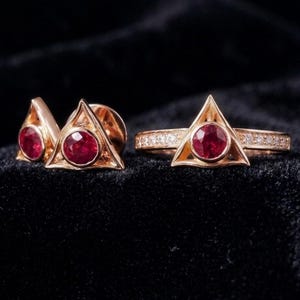 May include: A set of gold-colored jewelry featuring a ring and a pair of earrings, each with a triangular design. The ring has a band encrusted with small, clear stones. Each piece features a large, round, red gemstone at the center.