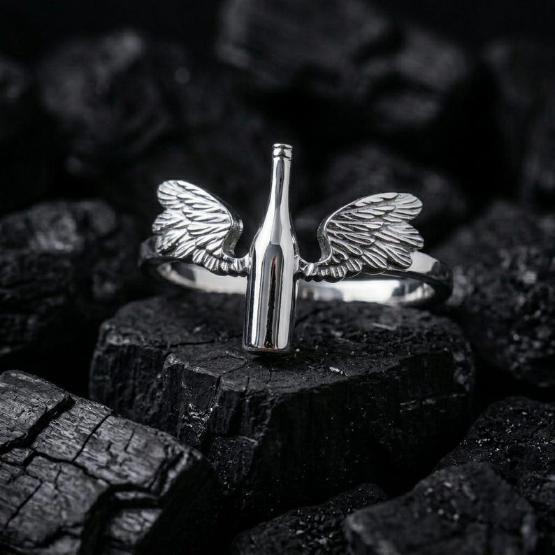 May include: A silver ring featuring a wine bottle design with detailed wings on either side. The ring is set against a dark, textured background, highlighting the metallic shine of the jewelry.