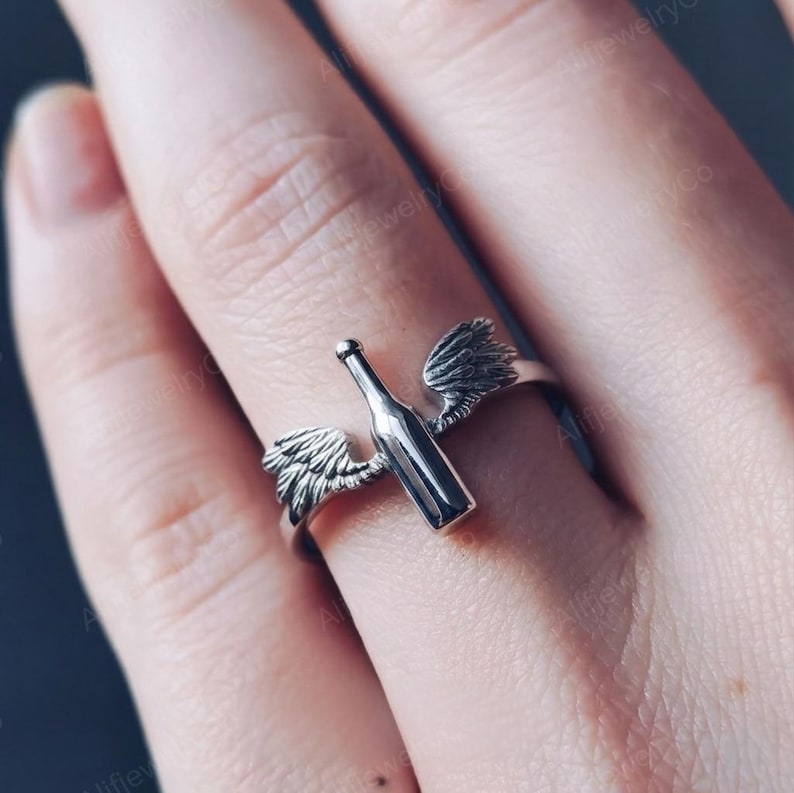 May include: A silver ring featuring a wine bottle design with feathered wings on either side. The ring is shown on a finger, with the bottle and wings extending across the top. The ring is made of a shiny metal.