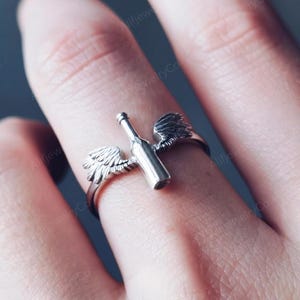 May include: A silver ring featuring a wine bottle design with two small wings on either side. The ring is made of a shiny metal and is shown on a finger. The ring's design is detailed and intricate.
