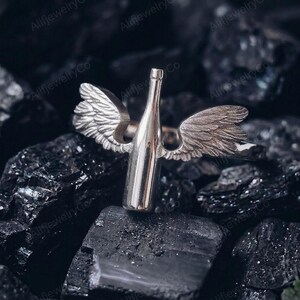 May include: A silver ring featuring a bottle design with angel wings. The ring is made of a shiny metal and is set against a dark, textured background. The wings are detailed, adding a unique touch to the jewelry.