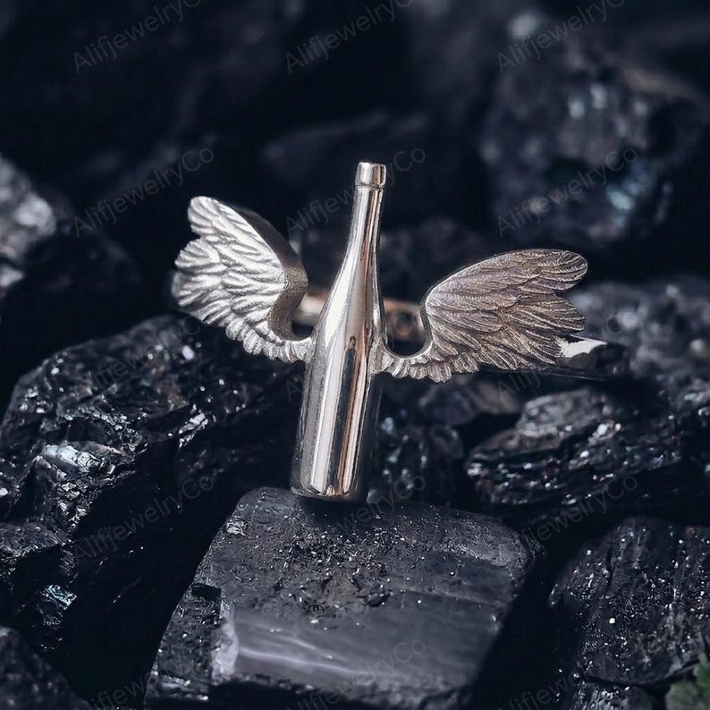 May include: A silver ring featuring a bottle design with angel wings. The ring is made of a shiny metal and is set against a dark, textured background. The wings are detailed, adding a unique touch to the jewelry.