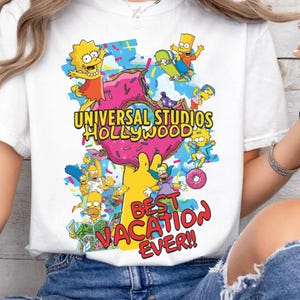 May include: White t-shirt featuring a colorful graphic of The Simpsons characters and the text "Universal Studios Hollywood". The design includes a large pink donut and the phrase "Best Vacation Ever!!".