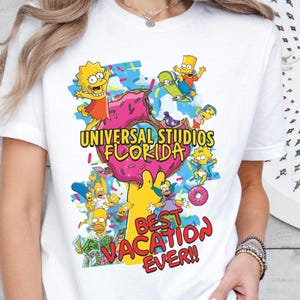 May include: White t-shirt featuring a colorful graphic of The Simpsons characters around a pink donut. The text reads "UNIVERSAL STUDIOS FLORIDA" and "BEST VACATION EVER!!". The design includes Bart, Lisa, Homer, and other characters.