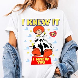 May include: A white t-shirt featuring a graphic of the character Jessie from Toy Story wearing a cow-print skirt and orange boots. The text reads I KNEW IT at the top and I KNEW YOU at the bottom, with a red heart containing the letters TS and yellow stars.