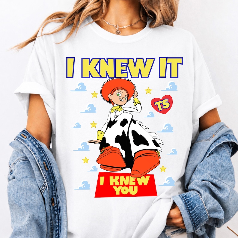 May include: A white t-shirt featuring a graphic of the character Jessie from Toy Story wearing a cow-print skirt and orange boots. The text reads I KNEW IT at the top and I KNEW YOU at the bottom, with a red heart containing the letters TS and yellow stars.