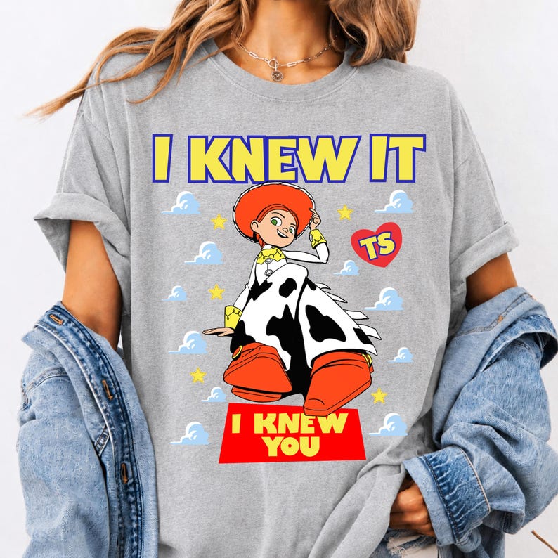 May include: A heather gray t-shirt featuring a graphic of the character Jessie from Toy Story. The text I KNEW IT appears at the top in yellow, and I KNEW YOU is printed in yellow on a red rectangle at the bottom. The design includes clouds and stars.
