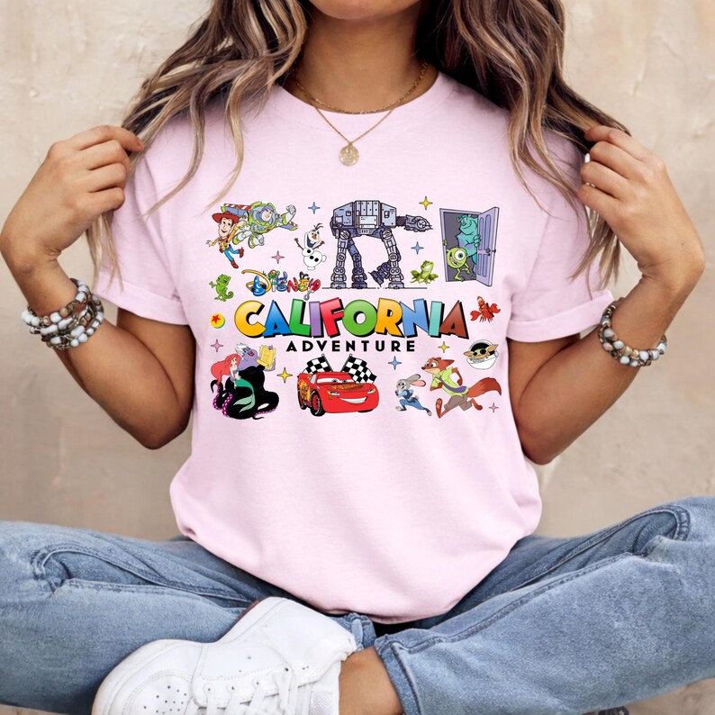 May include: A light pink t-shirt featuring colorful cartoon characters and the text "CALIFORNIA ADVENTURE". The design includes images of a red race car, an AT-AT walker, and other animated figures.
