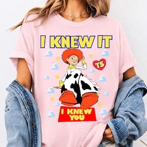 May include: A light pink t-shirt featuring a graphic of a cowgirl character with a red hat, white and black cow-print chaps, and orange boots. The text reads I KNEW IT at the top and I KNEW YOU at the bottom, with yellow stars and blue clouds.