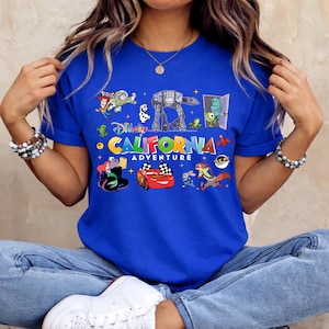 May include: Royal blue t-shirt featuring a colorful graphic with the text "CALIFORNIA ADVENTURE" and various cartoon characters. The design includes images of a race car, a monster, and other animated figures.