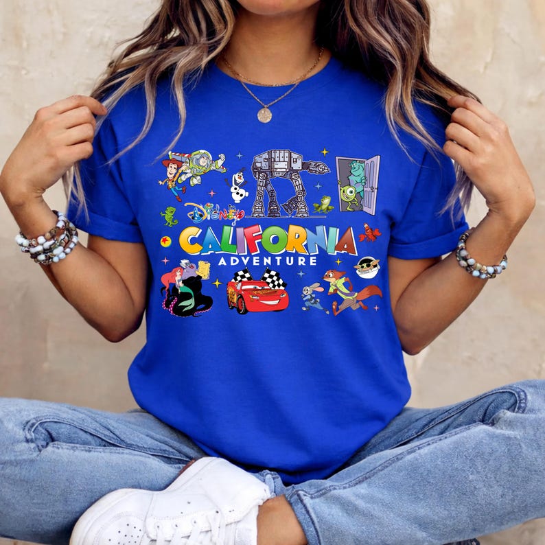 May include: Royal blue t-shirt featuring a colorful graphic with the text "CALIFORNIA ADVENTURE" and various cartoon characters. The design includes images of a race car, a monster, and other animated figures.